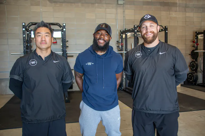 Three men standing in a gym setting, all wearing athletic gear, smiling for the camera. Weightlifting equipment is in the background.
