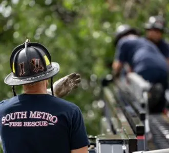 Firefighters from South Metro Fire Rescue on a ladder, performing a task amidst greenery.