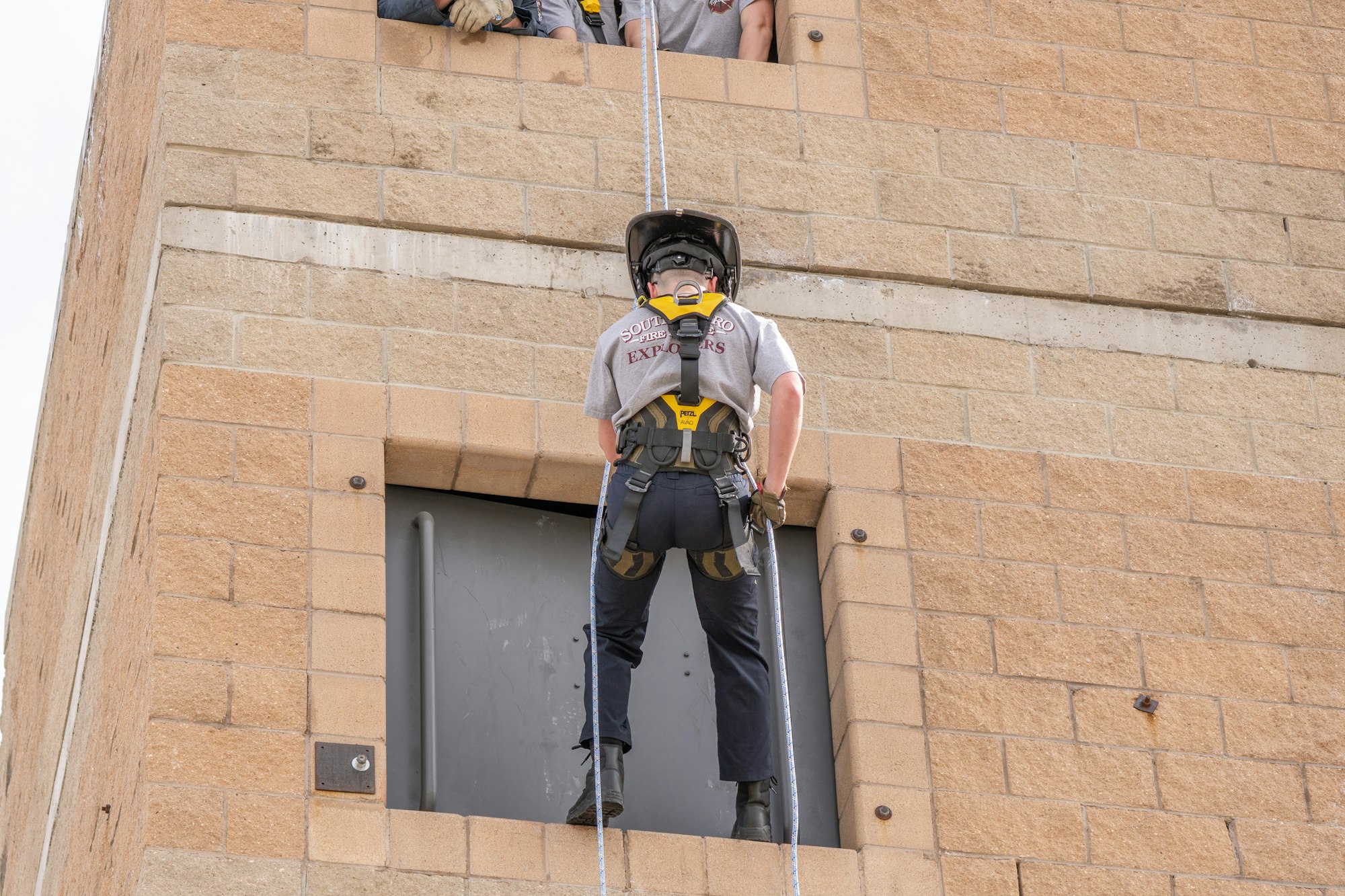 A person in safety gear is rappelling down a building, preparing to enter through a window while onlookers watch from above.