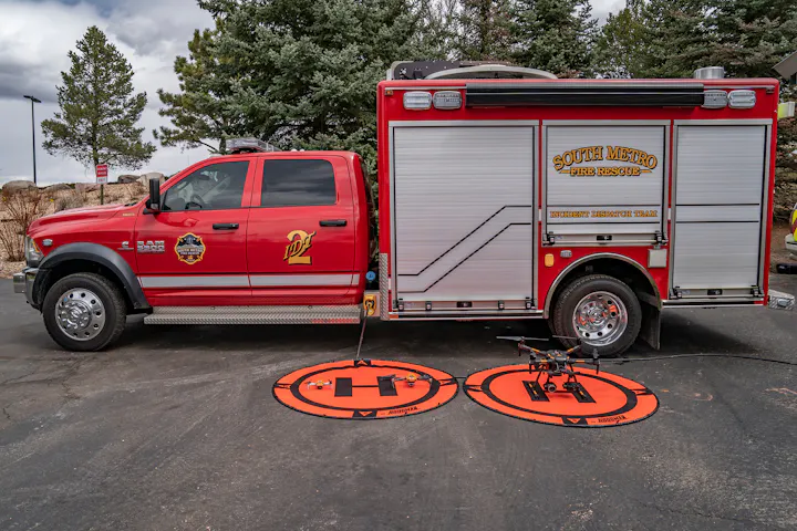 A red fire rescue truck parked with drone landing pads and a drone on the ground, set against a backdrop of trees and clouds.