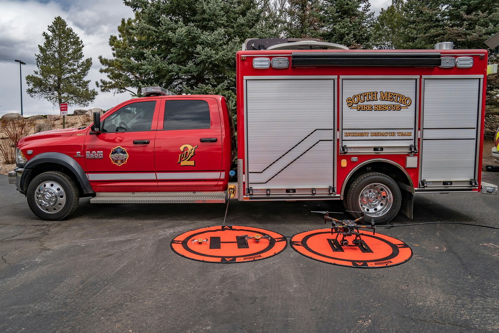 A red fire rescue truck parked with drone landing pads and a drone on the ground, set against a backdrop of trees and clouds.