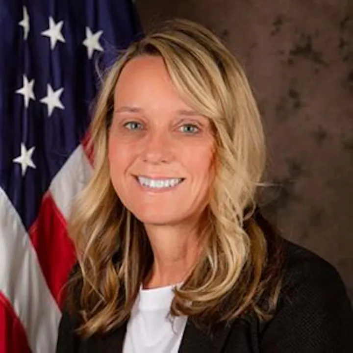 The image features a woman with blonde hair, smiling, standing in front of an American flag.
