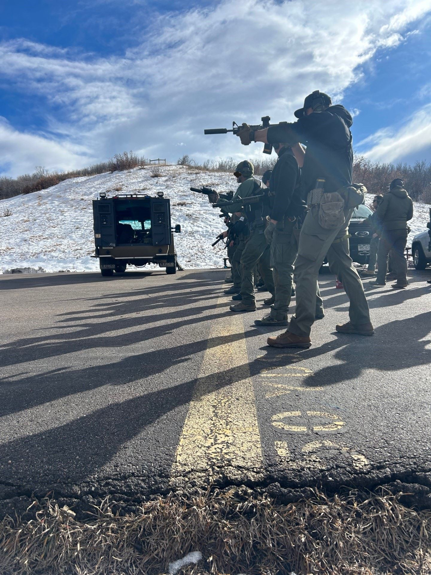 A group of people in tactical gear aiming firearms beside a snow-covered hill and an armored vehicle under a blue sky.