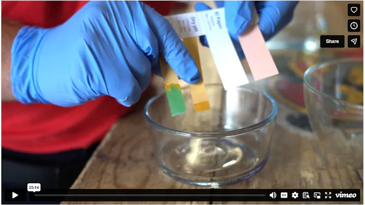 A person wearing gloves is holding pH test strips above a clear bowl on a wooden surface.