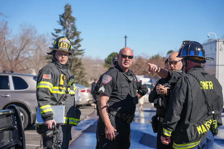 A firefighter and several police officers discuss something outdoors, with vehicles and trees in the background.