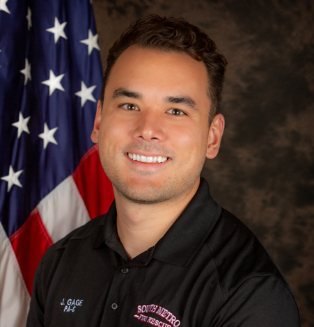 A smiling man in a black shirt stands in front of an American flag backdrop.