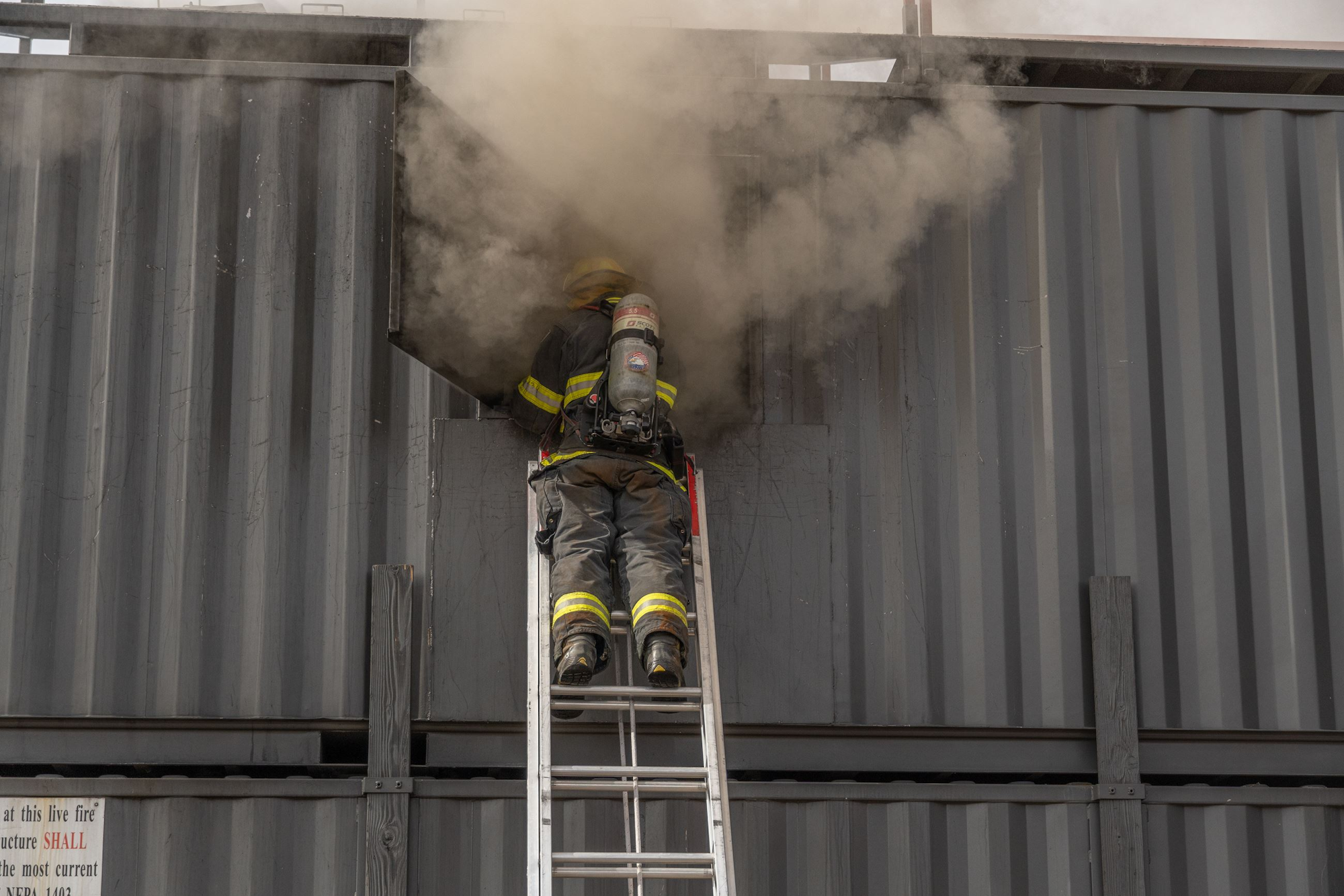 A firefighter on a ladder is battling smoke coming from an industrial container or structure. Safety gear is visible.