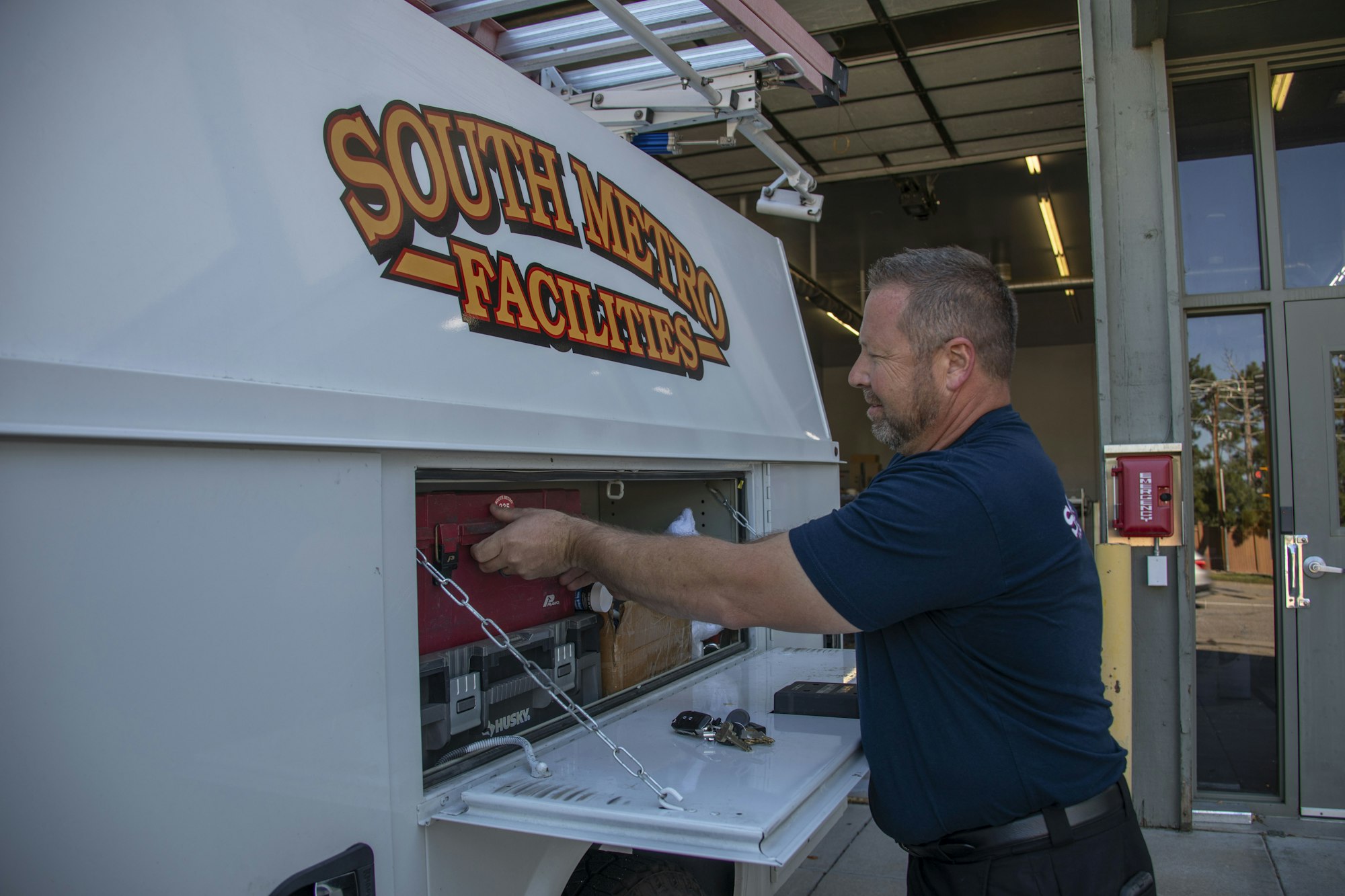 A man is opening a storage compartment on a vehicle marked "South Metro Facilities." Tools and equipment are inside.