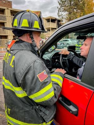 A firefighter in gear is talking to a person inside a red vehicle parked outdoors.