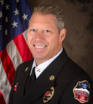 A person in a fire department uniform smiling, with the American flag in the background.
