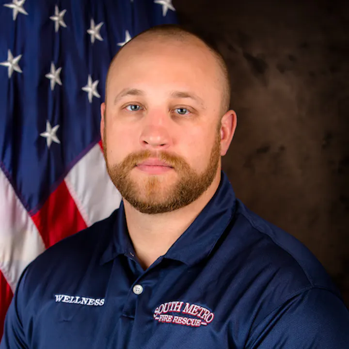The image shows a man in a navy blue shirt with "WELLNESS" and "SOUTH METRO FIRE RESCUE" logos, standing in front of an American flag.