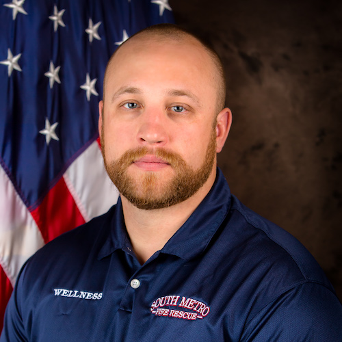 The image shows a man in a navy blue shirt with "WELLNESS" and "SOUTH METRO FIRE RESCUE" logos, standing in front of an American flag.