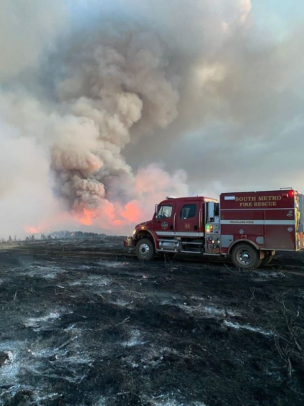 Fire truck in a burnt landscape, large smoke plume rising from a wildfire in the background.