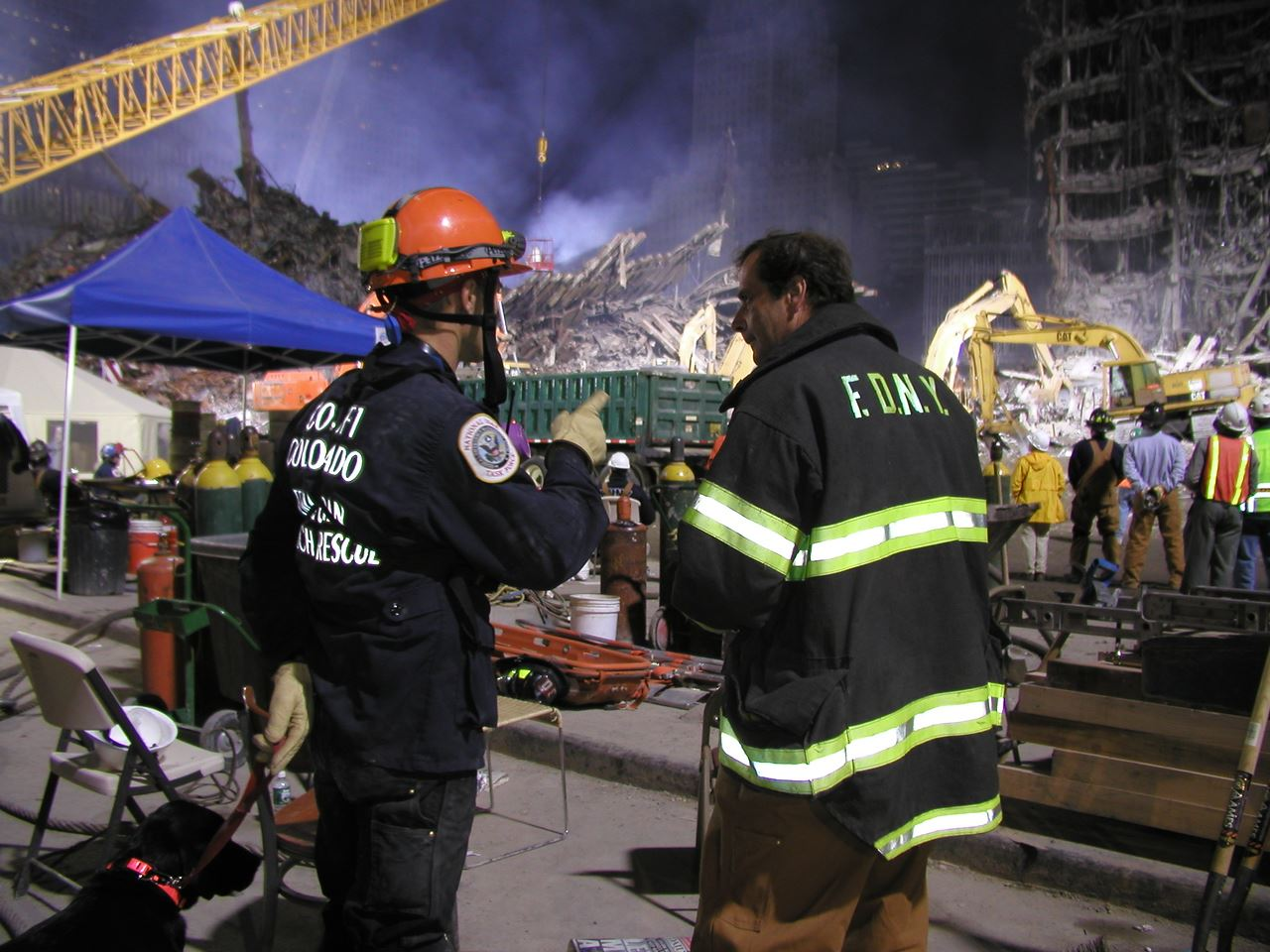 Rescue workers at a disaster site with debris, cranes, and equipment, discussing while others work nearby.