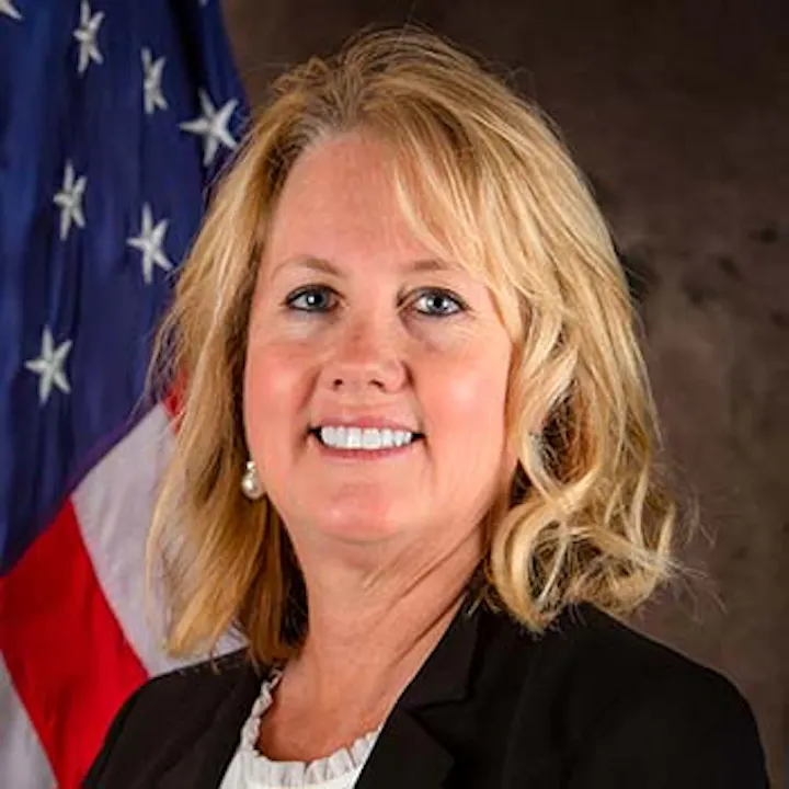 A smiling person with blonde hair is in front of a U.S. flag, wearing a black blazer and white top.