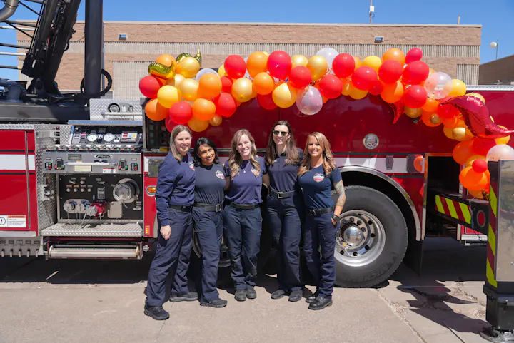 Five women in uniforms pose in front of a fire truck decorated with colorful balloons. It looks festive and celebratory.