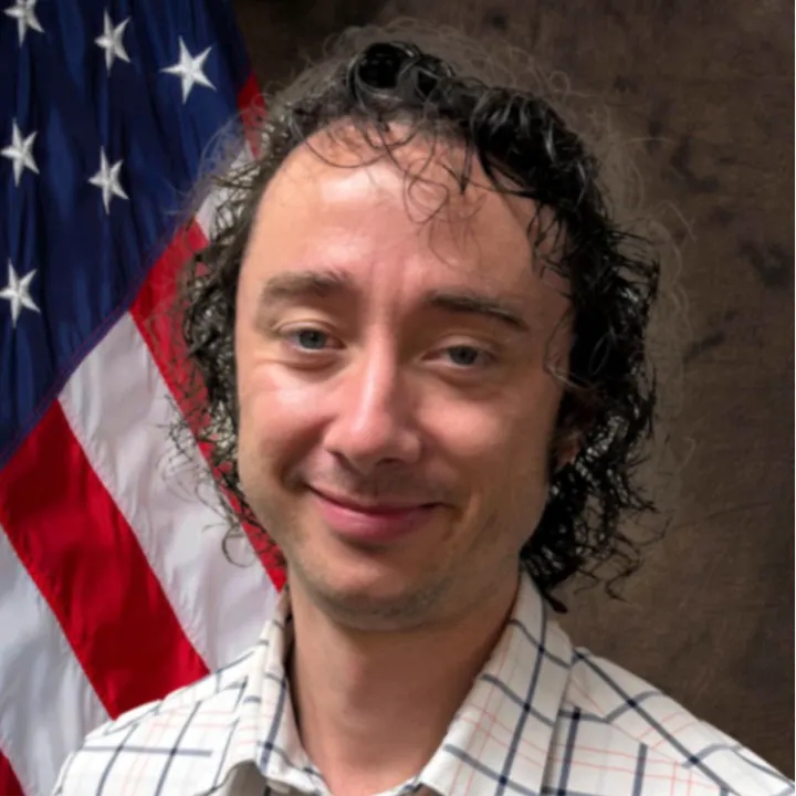 A person with curly hair and a plaid shirt in front of an American flag.