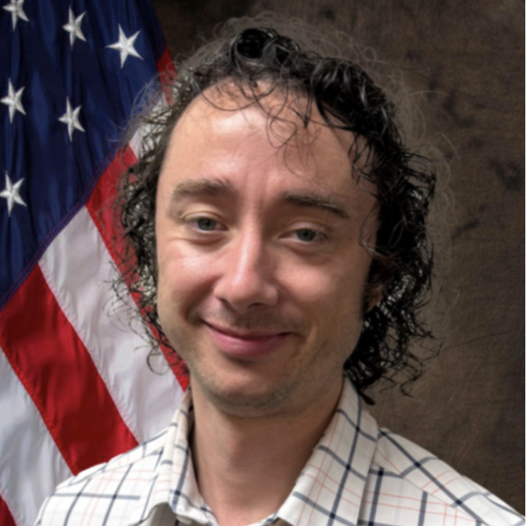 A person with curly hair and a plaid shirt in front of an American flag.