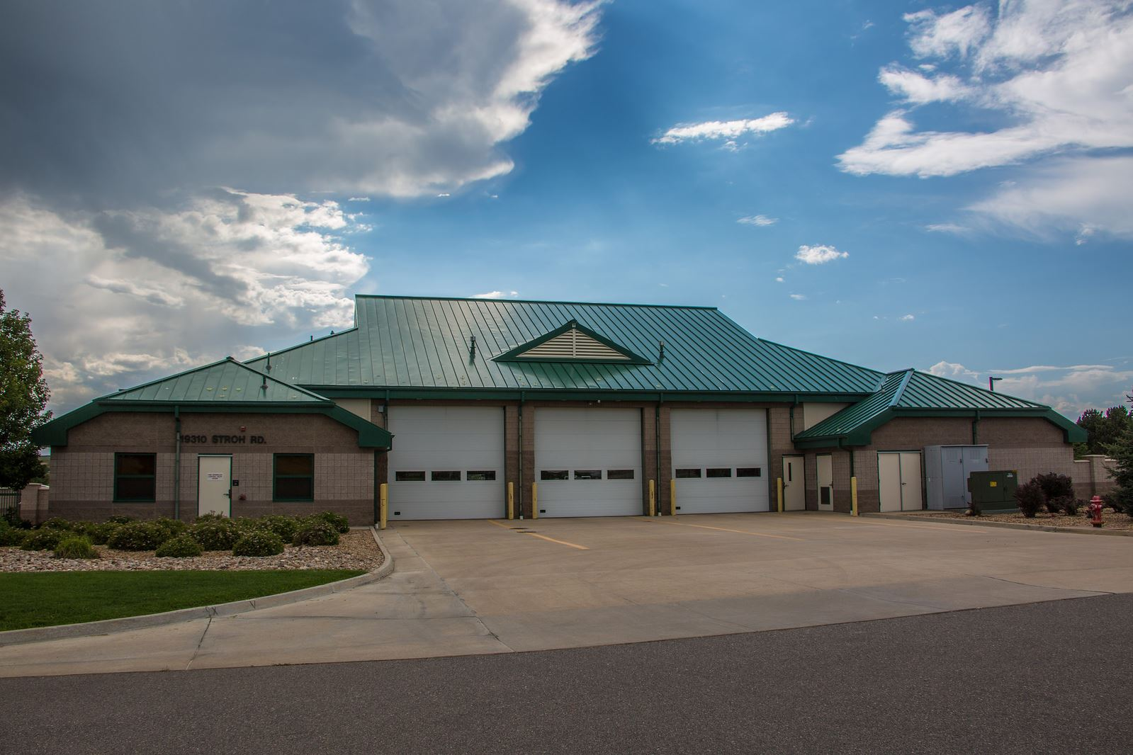 A building with a green roof and multiple garage doors, labeled "19310 Stroh Rd." under a partly cloudy sky.