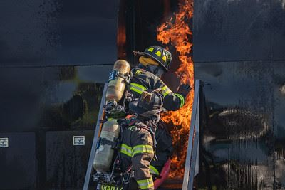 A firefighter enters a burning building, wearing full gear and carrying an oxygen tank. Flames are visible through the doorway.