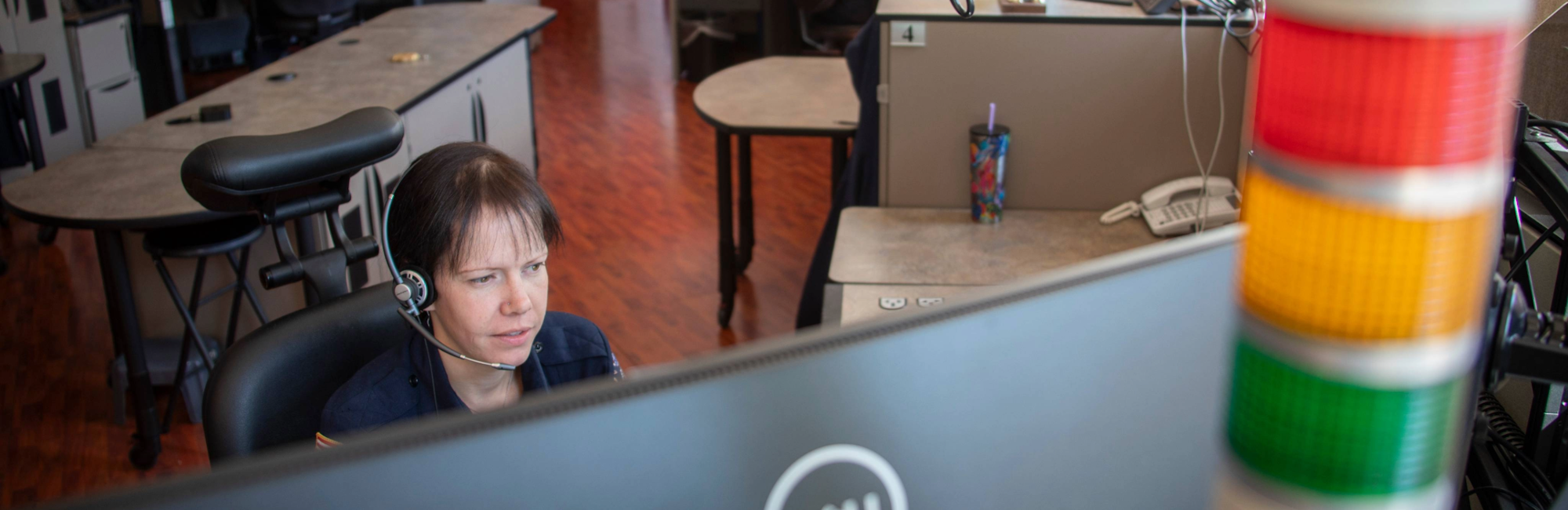 Person at a desk with a headset, large monitor, telephone, and a colorful cup; blurred control tower light in foreground.