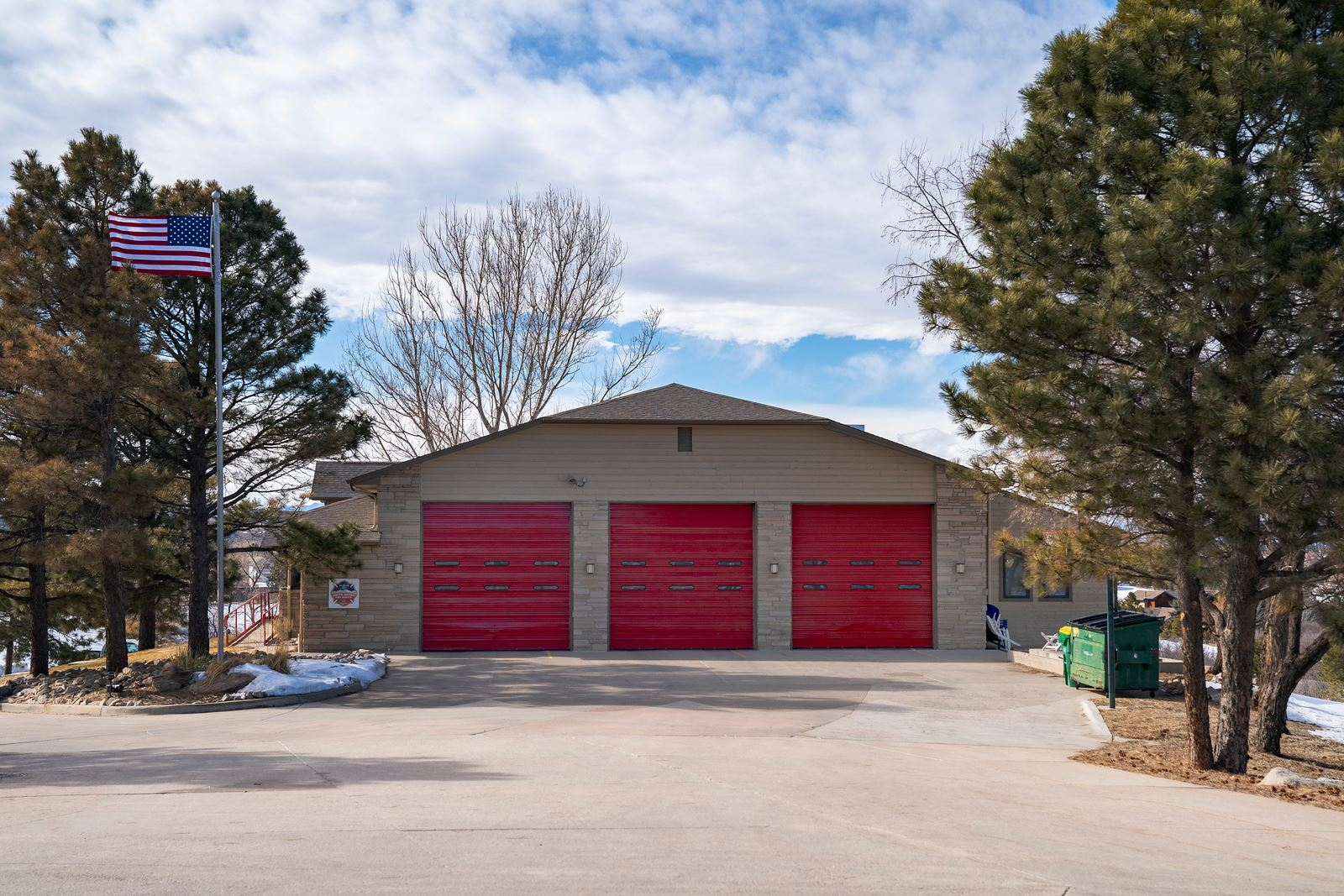 Building with three red garage doors, trees, an American flag, and a dumpster.