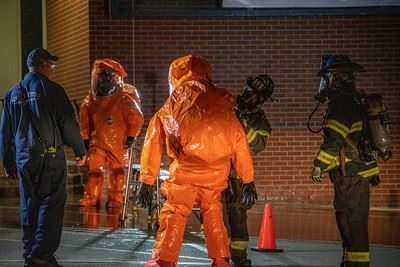 People in hazmat suits and firefighter gear are gathered near a brick wall with an orange traffic cone.