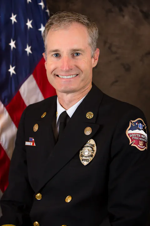 A smiling man in a uniform with badges, standing in front of an American flag.