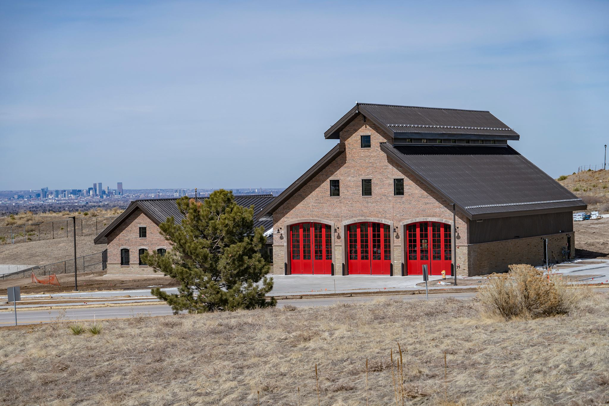 A brick building with red doors, surrounded by dry grass, with a city skyline visible in the distance.