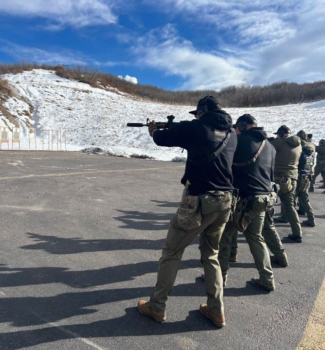 A group of people in tactical gear practicing shooting at an outdoor range with snowy hills in the background.