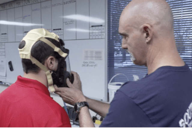 Two people in a room, one fitting a gas mask on the other, near a whiteboard.