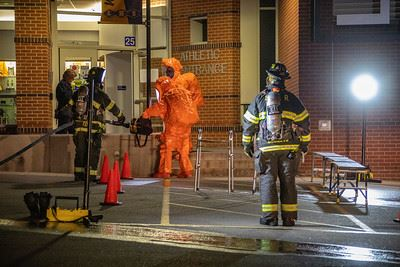 Firefighters in gear and a person in an orange hazmat suit are near an entrance, with equipment and cones set up outside.