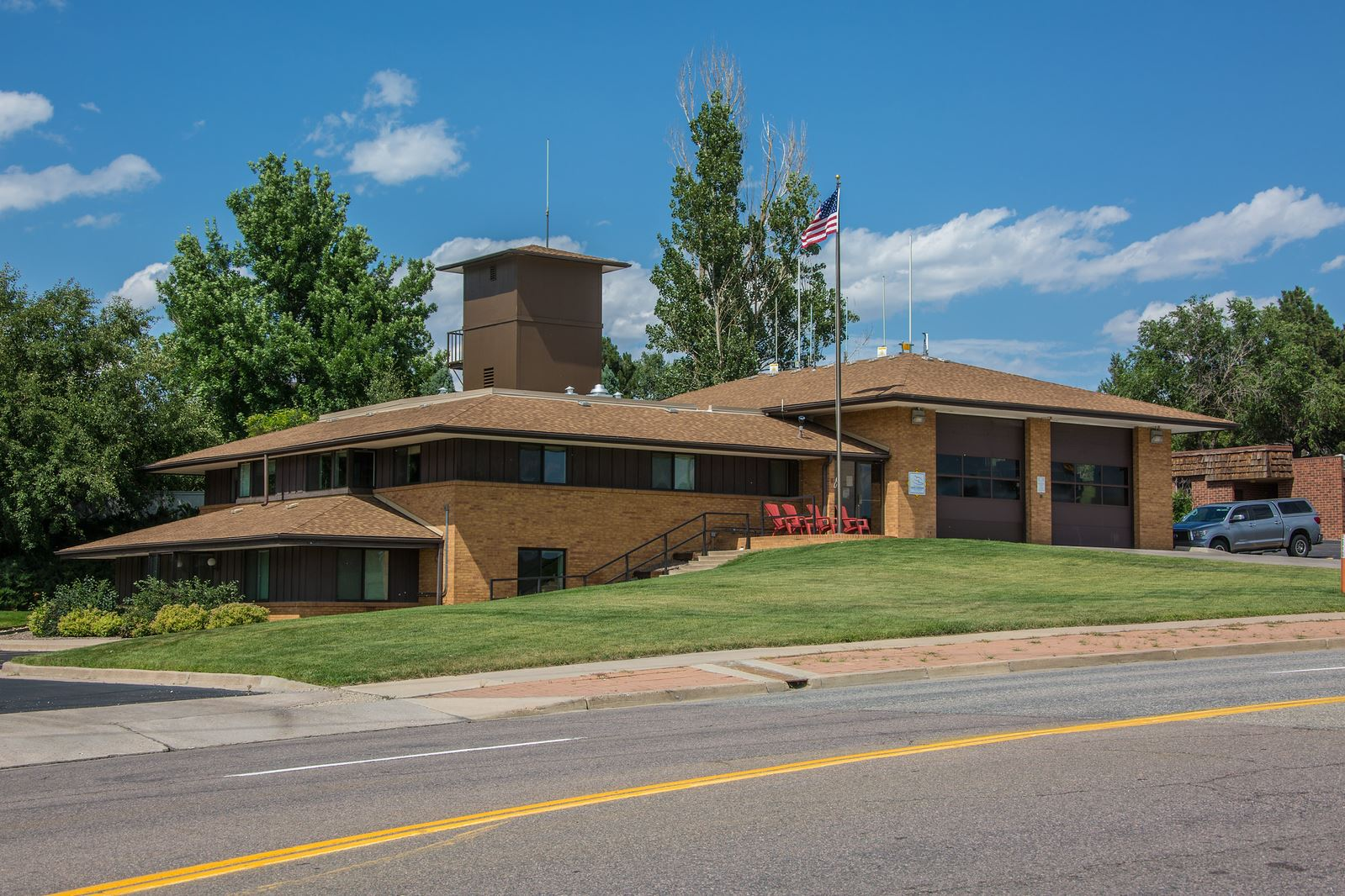 A brick building with a flagpole, surrounded by grass and trees, with a road in the foreground.