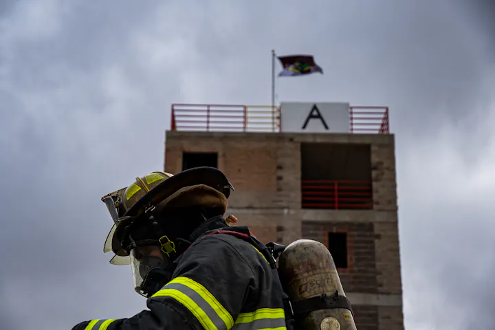 A firefighter in gear stands in front of a training tower, with a flag flying above. The sky looks overcast.