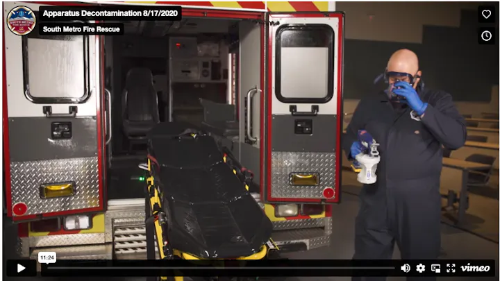 A firefighter in protective gear is preparing to decontaminate an ambulance with a spray bottle and a stretcher visible.