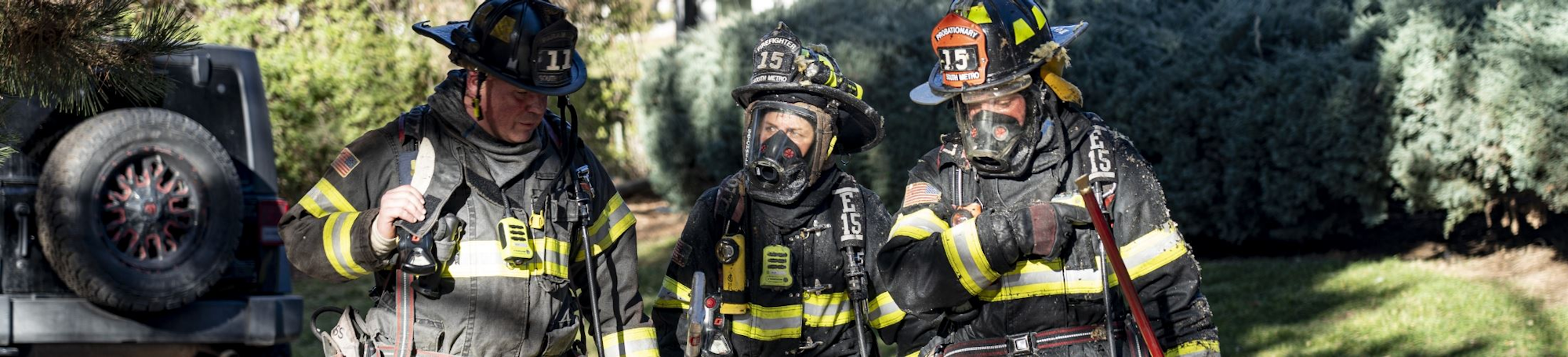 Three firefighters in gear stand outside, equipped with helmets and breathing masks, next to a vehicle with a spare tire.