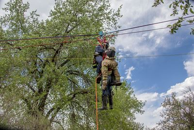Two people are suspended in the air, climbing a rope near trees under a partly cloudy sky.