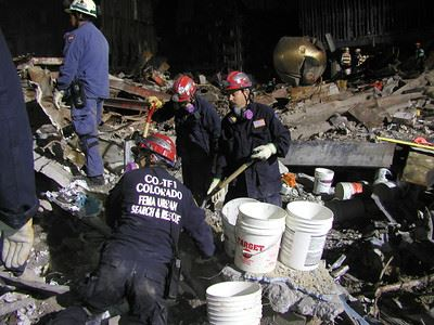Rescue workers in uniforms and helmets search through rubble with buckets and tools, likely in a disaster site.