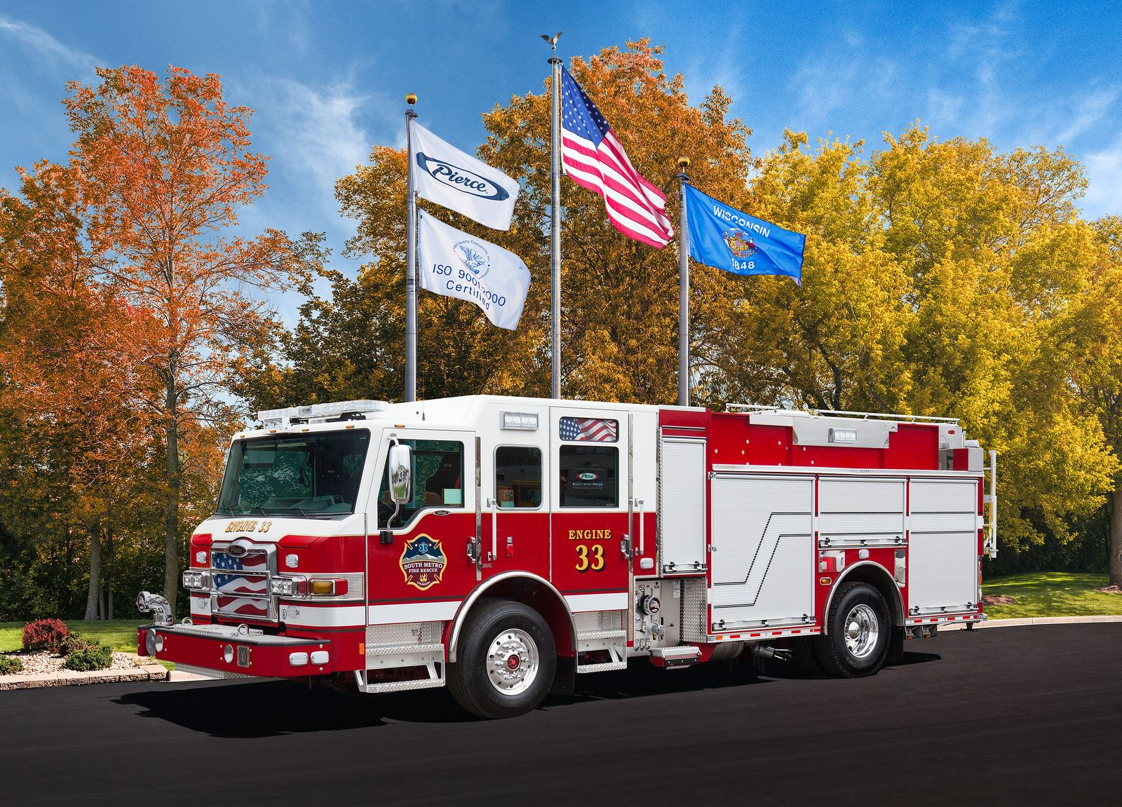 A red and white fire truck, labeled "Engine 33," with flags in the background, parked on a road with autumn trees.