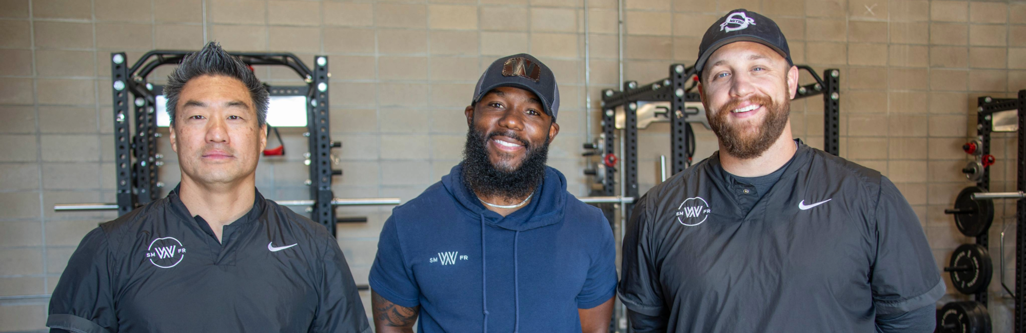 Three people smiling in a gym, wearing athletic clothing and caps.