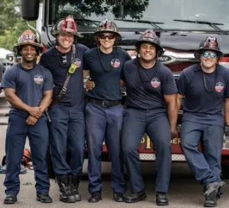 Five firefighters in uniform smiling in front of a fire truck.