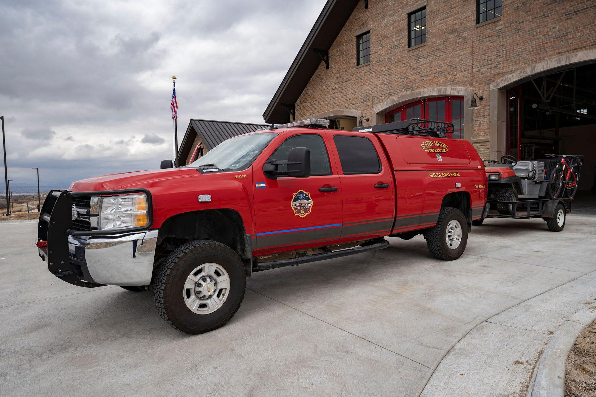 Red fire truck with a trailer carrying equipment, parked outside a brick fire station with an American flag nearby.