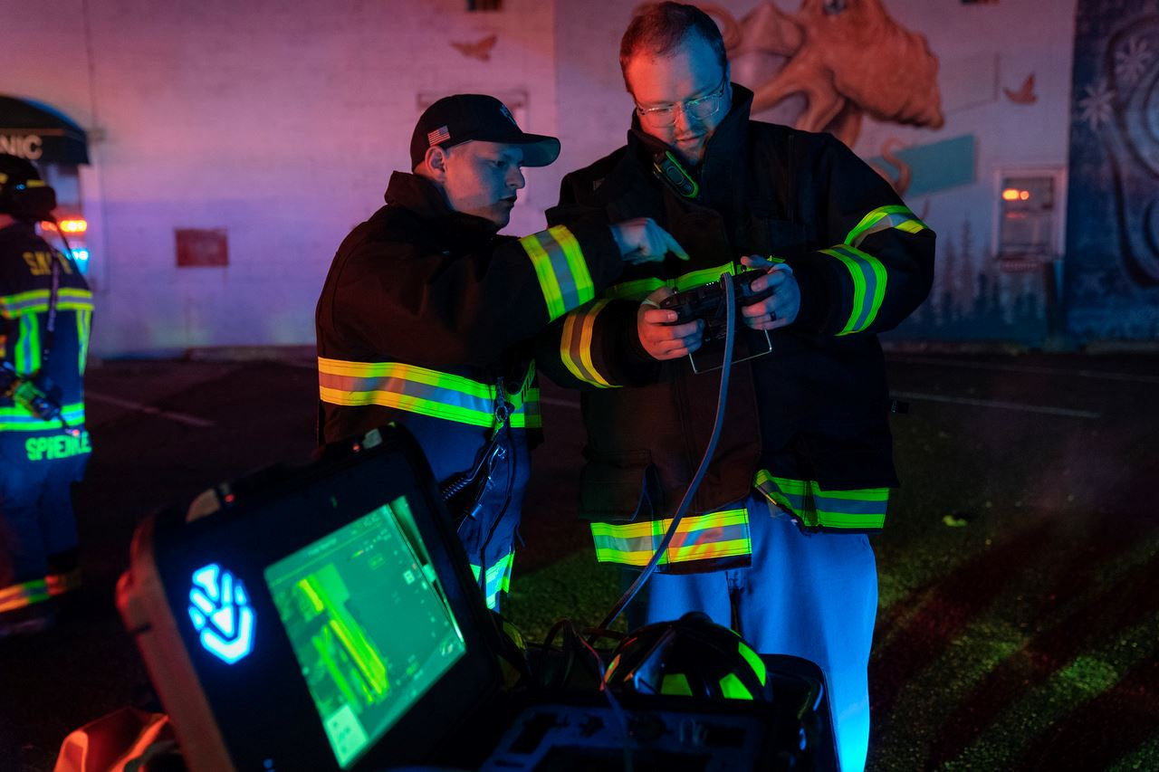 Firefighters operating a device with a screen showing thermal imaging in a dimly lit area.