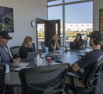A group of five people having a meeting around a table with laptops, papers, and drinks in a modern office setting.