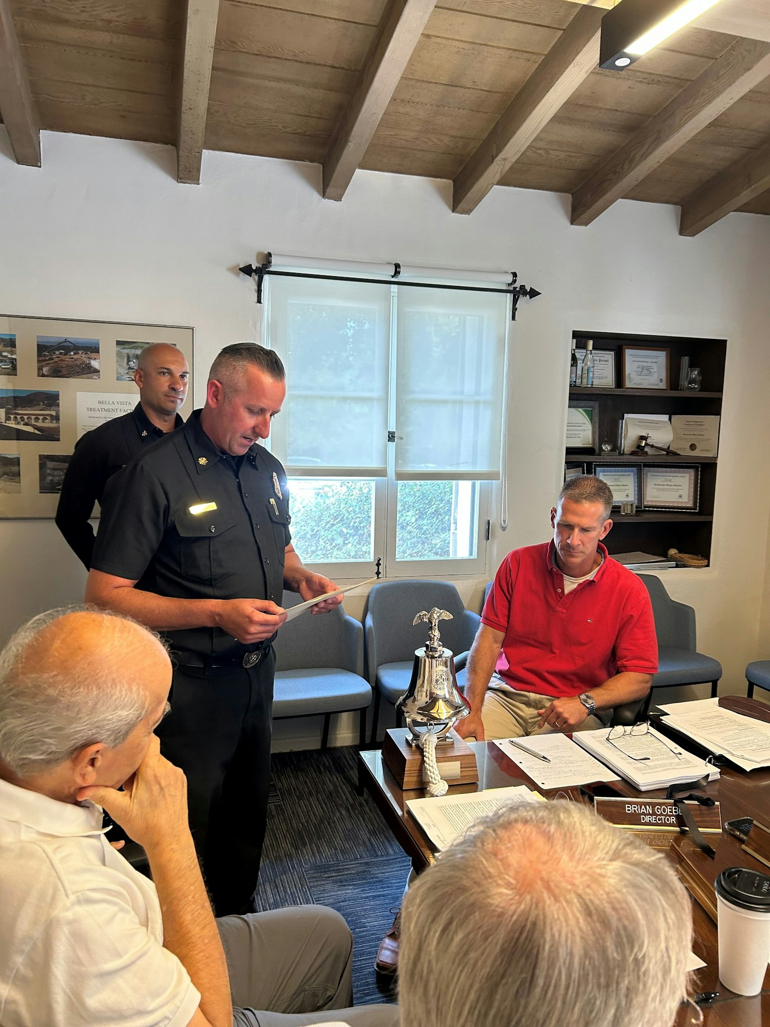 A police officer is speaking to a group during a meeting, with documents on the table and a decorative bell in the foreground.