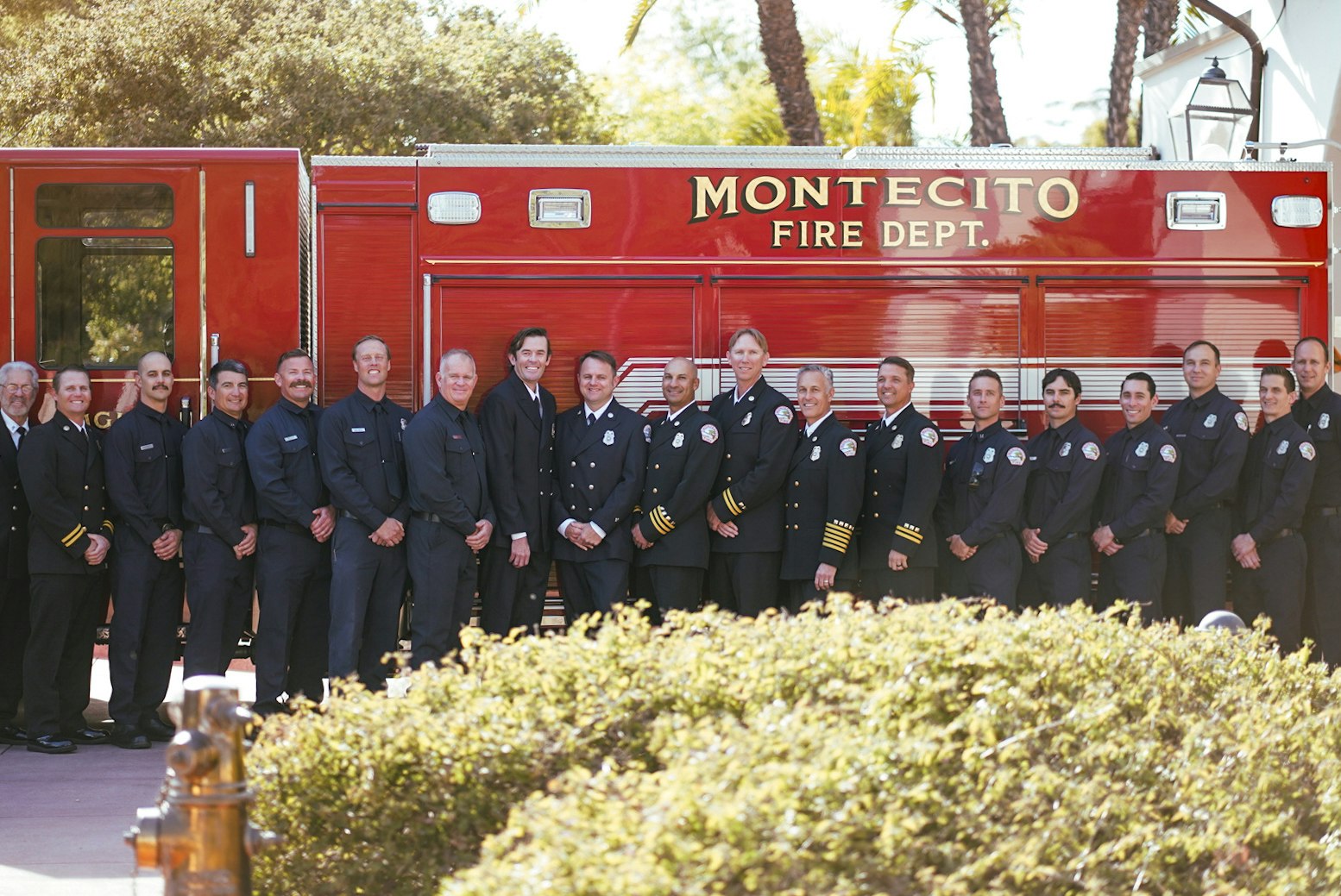 A group of firefighters in uniform standing in front of a Montecito Fire Dept. fire truck, with trees and bushes in the background.