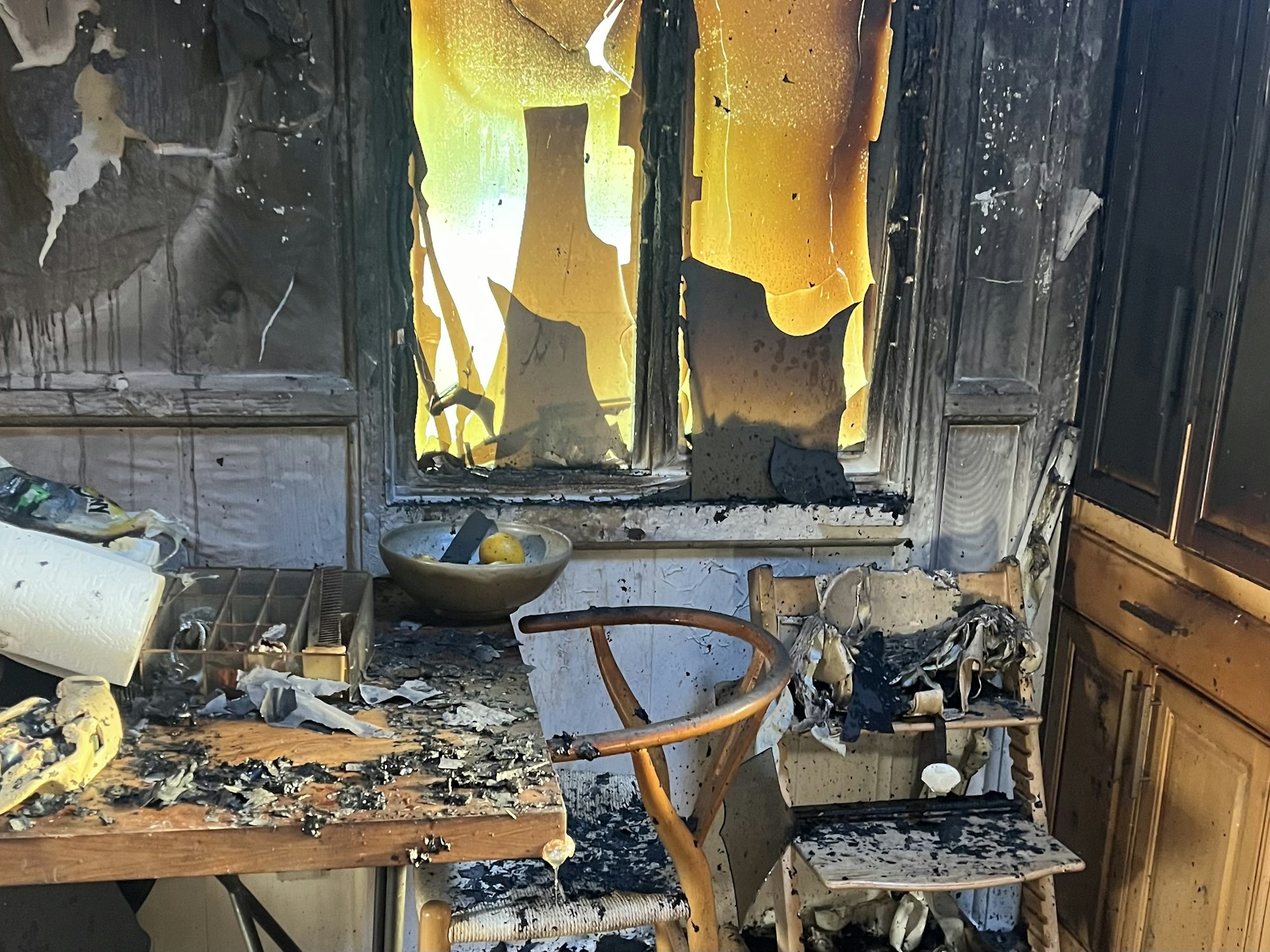 A burnt room with charred furniture, melted window blinds, and scattered debris.