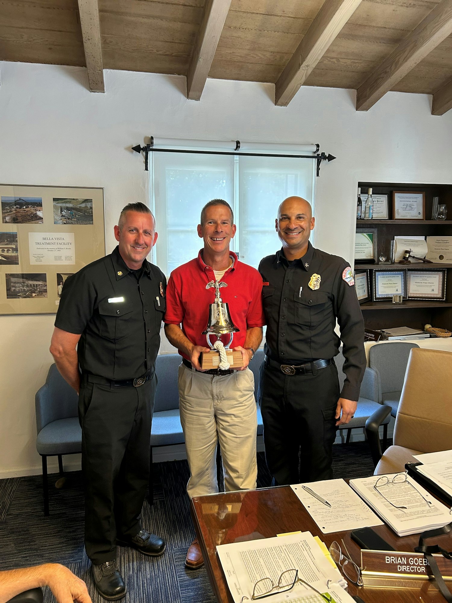 Three men, two in uniforms and one in a red shirt, are smiling while holding a decorative bell trophy in an office setting.