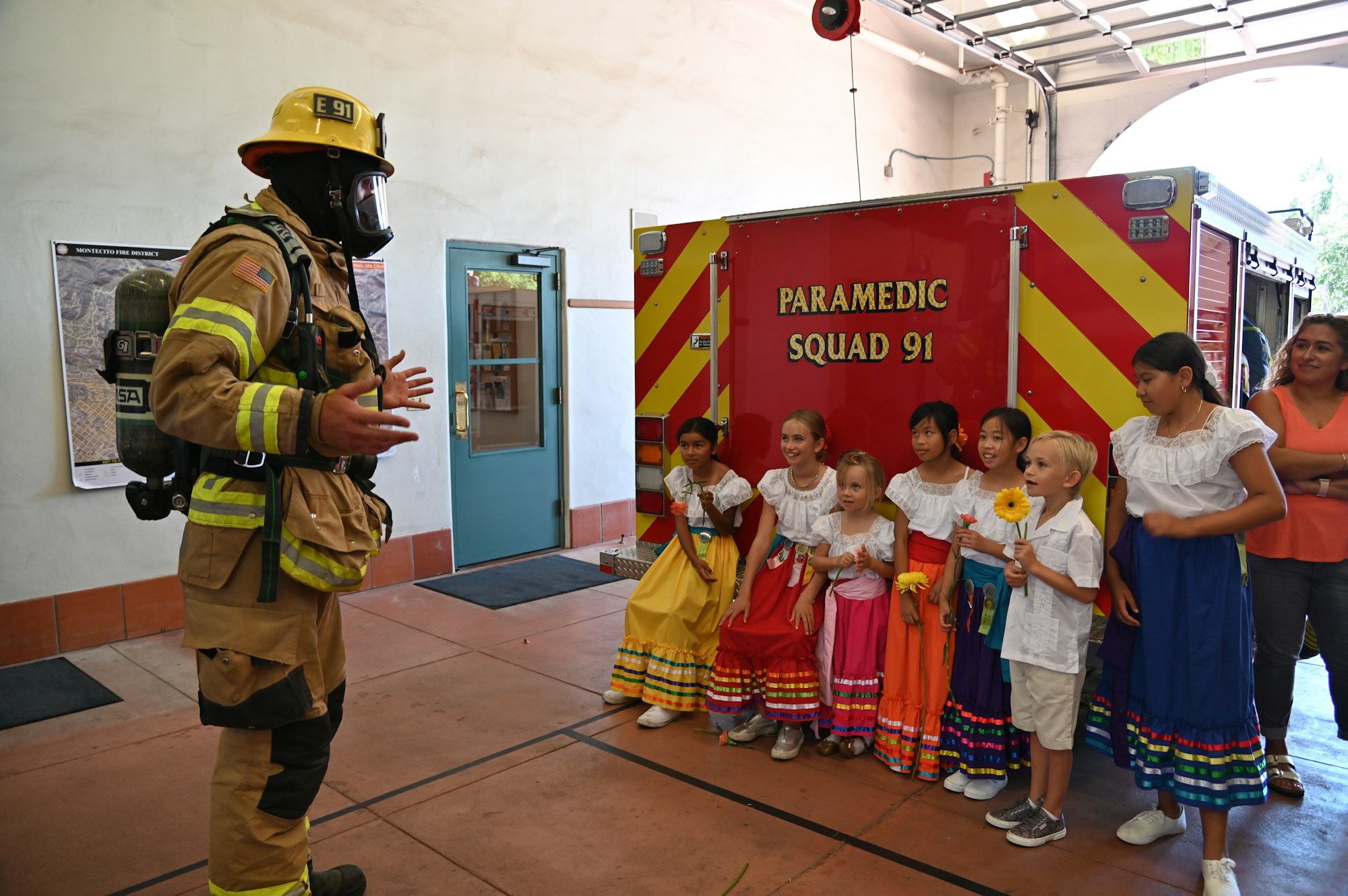 A firefighter is presenting to children in cultural dresses beside a paramedic vehicle.