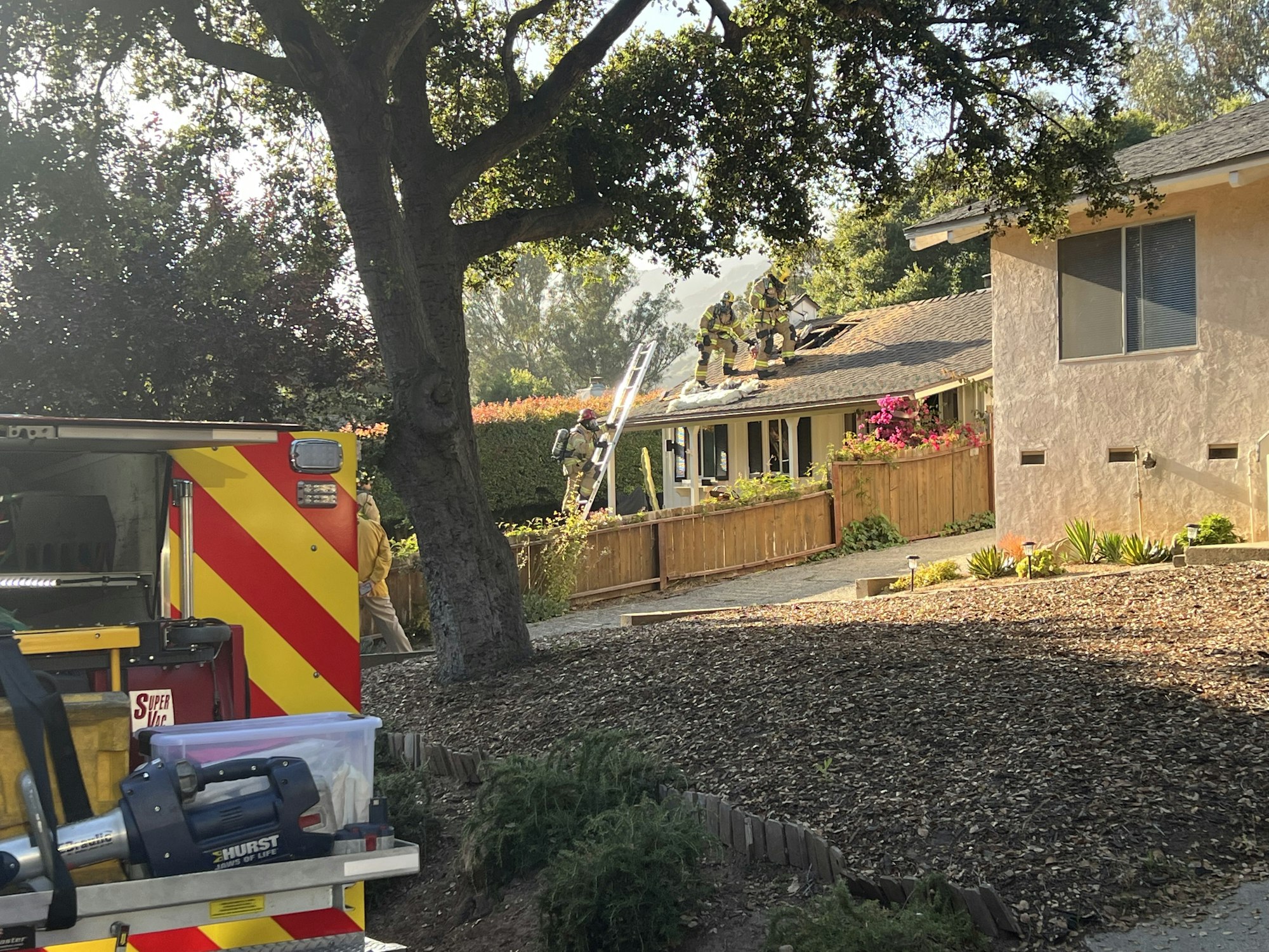 Firefighters on a house roof, fire truck in the foreground, in a residential area with trees and sunlight.